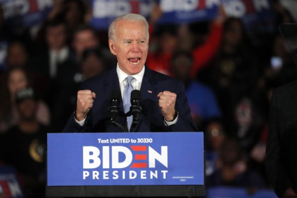 Biden speaking to a crowd behind a podium reading 'BIDEN PRESIDENT'